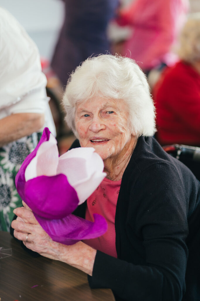 Beryl at Southern Cross Care WA East Fremantle is smiling and holding a large purple and pink crepe paper flower she has made on International Nurse Day.